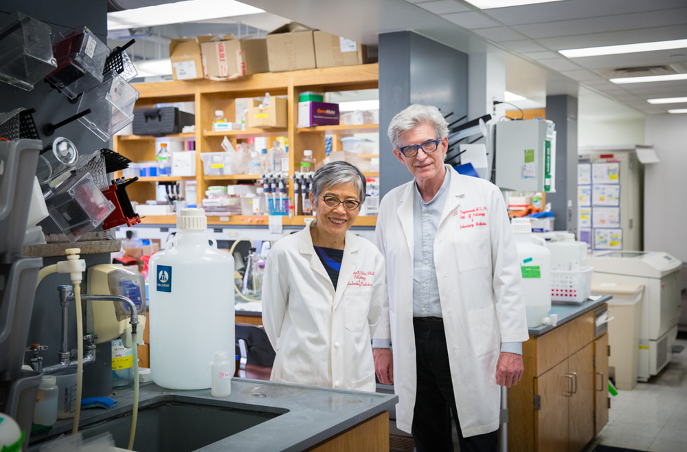 Virginia Lee, PhD, and John Trojanowski, MD, PhD, wearing white coats in a lab in 2018 or 2019. 